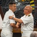 Two naval officers in white dress uniforms are engaged in what appears to be a formal military ceremony, likely a promotion or award pinning. The officer on the right, who has gold shoulder boards indicating senior rank, is placing or adjusting something on the shoulder or collar of the officer on the left. The setting appears to be an indoor military chapel or ceremonial hall with a decorative mosaic or stained glass backdrop.