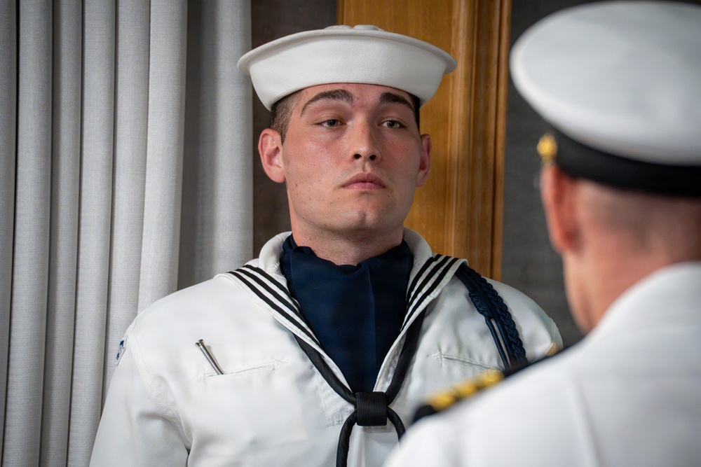 A young naval enlisted sailor in dress white uniform stands at attention while facing a senior officer whose back is to the camera. The sailor is wearing a traditional 'Crackerjack' dress white uniform with a navy blue neckerchief, white dixie cup hat, and a blue aiguillette, suggesting a ceremonial or formal inspection context. The setting appears to be an interior formal space with columns and wood paneling visible in the background.