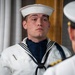 A young naval enlisted sailor in dress white uniform stands at attention while facing a senior officer whose back is to the camera. The sailor is wearing a traditional 'Crackerjack' dress white uniform with a navy blue neckerchief, white dixie cup hat, and a blue aiguillette, suggesting a ceremonial or formal inspection context. The setting appears to be an interior formal space with columns and wood paneling visible in the background.