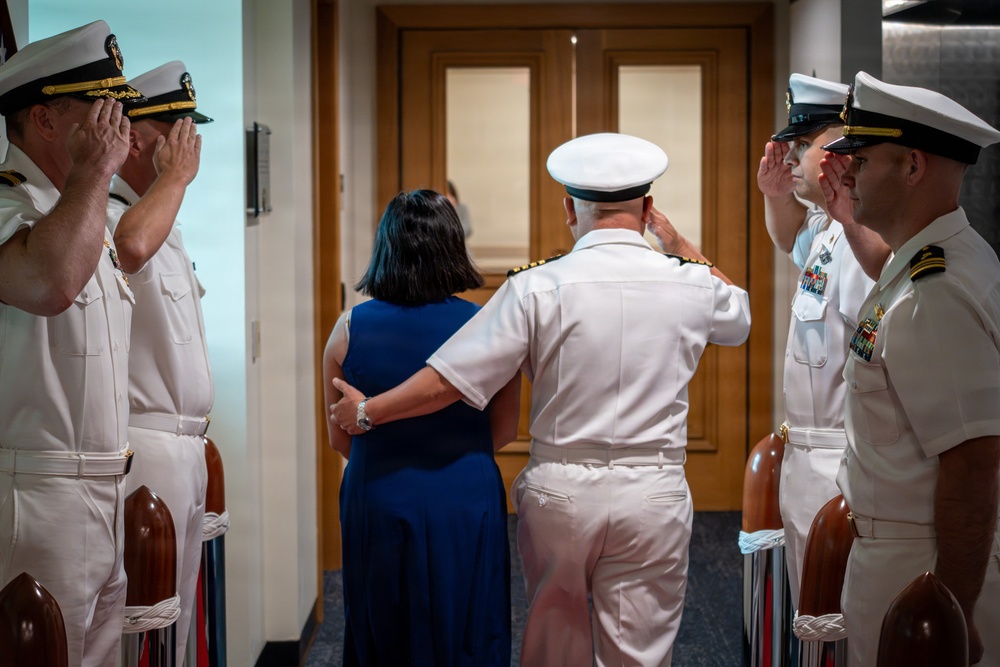 A formal U.S. Navy military ceremony is taking place, likely a change of command or retirement ceremony. A senior naval officer in white dress uniform escorts a civilian woman (presumably his spouse) through a sideboy honor guard. Four uniformed naval officers are rendered salutes as the couple passes through the corridor.