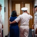 A formal U.S. Navy military ceremony is taking place, likely a change of command or retirement ceremony. A senior naval officer in white dress uniform escorts a civilian woman (presumably his spouse) through a sideboy honor guard. Four uniformed naval officers are rendered salutes as the couple passes through the corridor.