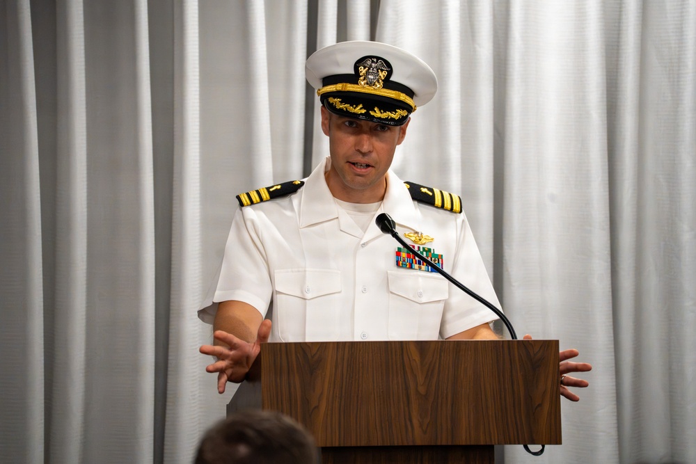 A U.S. Navy officer in white service dress uniform is delivering a speech at a wooden podium. The officer appears to be of senior rank, indicated by gold sleeve stripes and command-level cap insignia. The setting appears to be a formal indoor ceremony or briefing.
