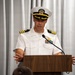 A U.S. Navy officer in white service dress uniform is delivering a speech at a wooden podium. The officer appears to be of senior rank, indicated by gold sleeve stripes and command-level cap insignia. The setting appears to be a formal indoor ceremony or briefing.