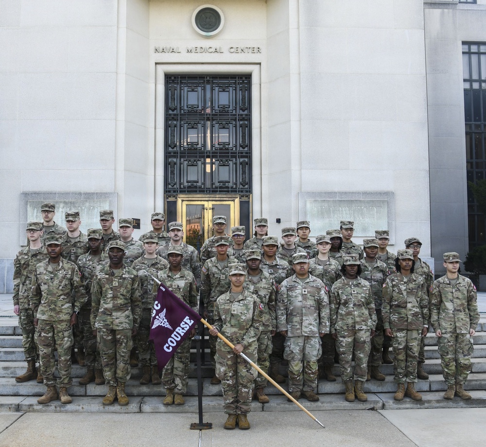 A large group of military personnel in U.S. Army camouflage (OCP/MultiCam) uniforms pose for a formal group photograph on the steps of the Naval Medical Center. A soldier in the front center holds a maroon/burgundy guidon flag with a caduceus symbol and the word 'COURSE' visible. The setting suggests this is a graduation or class photo for a military medical training course.