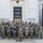 A large group of military personnel in U.S. Army camouflage (OCP/MultiCam) uniforms pose for a formal group photograph on the steps of the Naval Medical Center. A soldier in the front center holds a maroon/burgundy guidon flag with a caduceus symbol and the word 'COURSE' visible. The setting suggests this is a graduation or class photo for a military medical training course.
