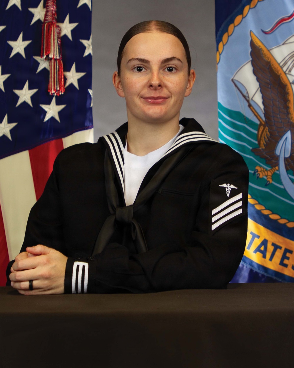 A formal military portrait of a U.S. Navy enlisted sailor in dress blue uniform, seated at a table with hands clasped. The subject wears a rating badge indicating a Hospital Corpsman (HM) petty officer third class rank. Two flags are visible in the background: the American flag and the United States Navy flag.