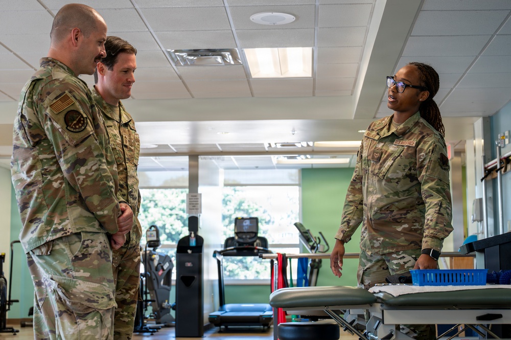 Three U.S. Air Force personnel in OCP (Operational Camouflage Pattern) uniforms are engaged in conversation inside what appears to be a military physical therapy or rehabilitation facility. A female Airman on the right, wearing glasses and identified by 'U.S. AIR FORCE' tape on her uniform, appears to be briefing or explaining something to two male service members. The setting includes exercise equipment and a treatment table in the background.