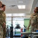 Three U.S. Air Force personnel in OCP (Operational Camouflage Pattern) uniforms are engaged in conversation inside what appears to be a military physical therapy or rehabilitation facility. A female Airman on the right, wearing glasses and identified by 'U.S. AIR FORCE' tape on her uniform, appears to be briefing or explaining something to two male service members. The setting includes exercise equipment and a treatment table in the background.
