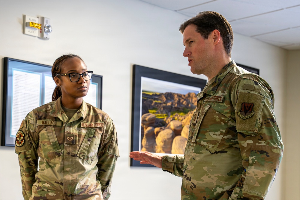 Two U.S. Air Force personnel in OCP (Operational Camouflage Pattern) uniforms are engaged in conversation inside what appears to be a military facility. The individual on the left, whose name tape reads 'WALLACE,' is listening attentively while the individual on the right appears to be speaking and gesturing with their hand. Both wear standard U.S. Air Force branch tape and unit patches on their sleeves.