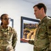 Two U.S. Air Force personnel in OCP (Operational Camouflage Pattern) uniforms are engaged in conversation inside what appears to be a military facility. The individual on the left, whose name tape reads 'WALLACE,' is listening attentively while the individual on the right appears to be speaking and gesturing with their hand. Both wear standard U.S. Air Force branch tape and unit patches on their sleeves.