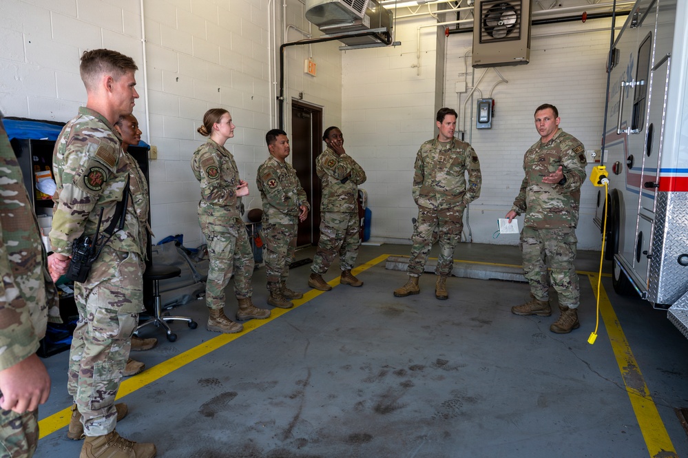A group of approximately seven military personnel in OCP (Operational Camouflage Pattern) uniforms are gathered inside what appears to be an emergency vehicle bay or fire station garage. One individual on the right appears to be briefing or instructing the group, holding papers, while standing near the rear of an ambulance. The setting suggests a training session, shift briefing, or operational readiness review.