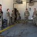 A group of approximately seven military personnel in OCP (Operational Camouflage Pattern) uniforms are gathered inside what appears to be an emergency vehicle bay or fire station garage. One individual on the right appears to be briefing or instructing the group, holding papers, while standing near the rear of an ambulance. The setting suggests a training session, shift briefing, or operational readiness review.