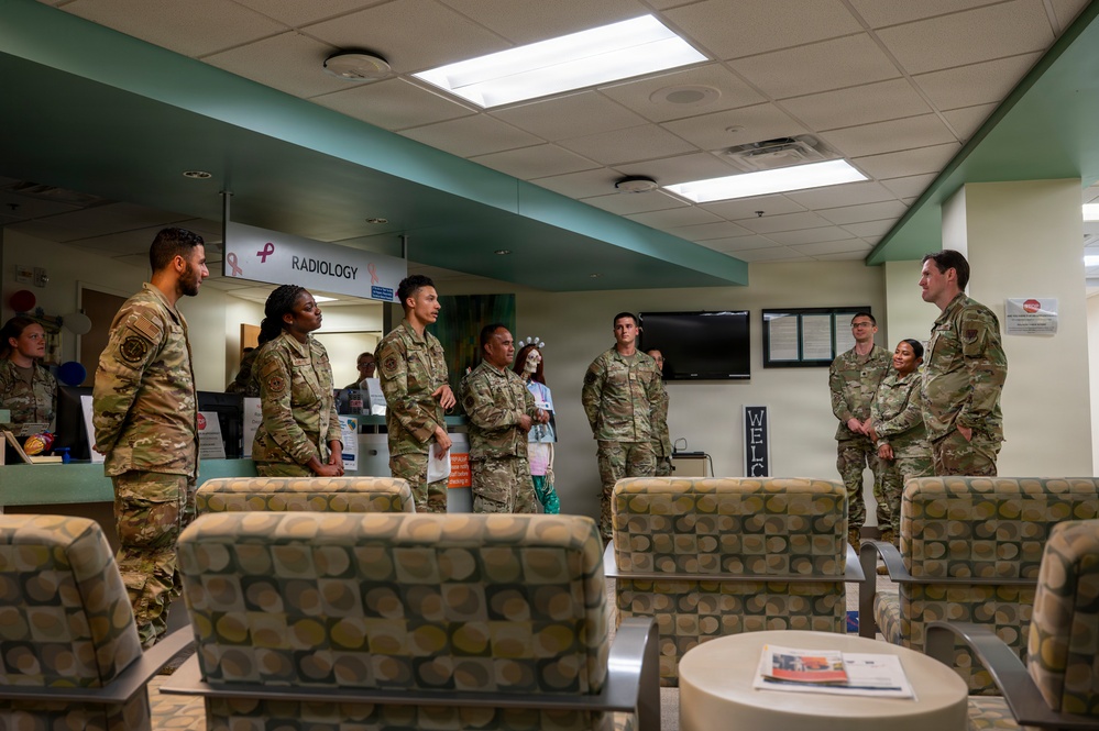 A group of military personnel in OCP (Operational Camouflage Pattern) uniforms are gathered in a medical facility waiting area near a Radiology department. They appear to be receiving a briefing or tour, with one individual on the right appearing to address the group. The setting is a clinical waiting room with patterned seating and standard medical facility decor.