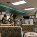 A group of military personnel in OCP (Operational Camouflage Pattern) uniforms are gathered in a medical facility waiting area near a Radiology department. They appear to be receiving a briefing or tour, with one individual on the right appearing to address the group. The setting is a clinical waiting room with patterned seating and standard medical facility decor.