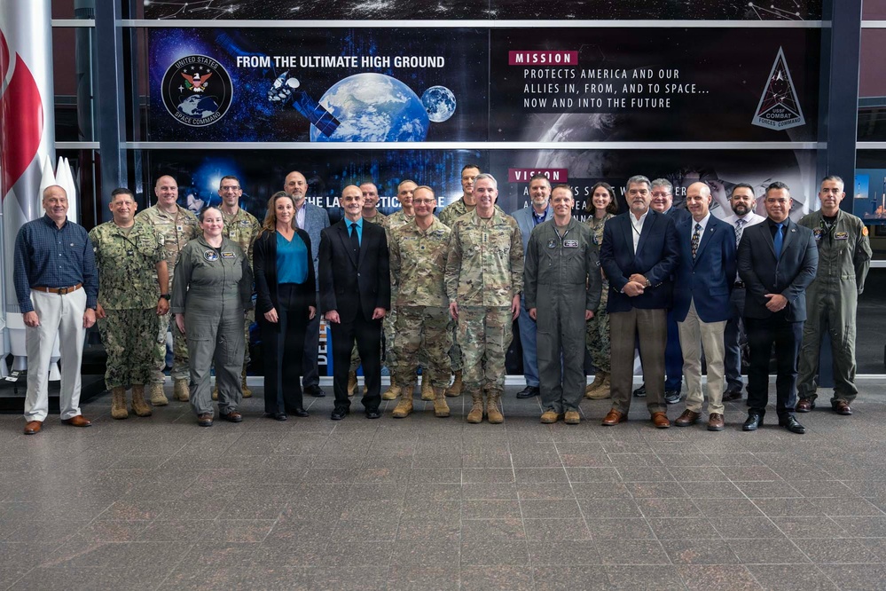 A group of approximately 20 military personnel and civilians pose for a formal group photograph inside a United States Space Command facility. The backdrop features official Space Command signage displaying the organization's mission statement 'Protects America and our allies in, from, and to space... now and into the future' and the tagline 'From the Ultimate High Ground.' The group includes individuals in various military uniforms (Army, Navy, Space Force, and flight suits) as well as civilian attire.