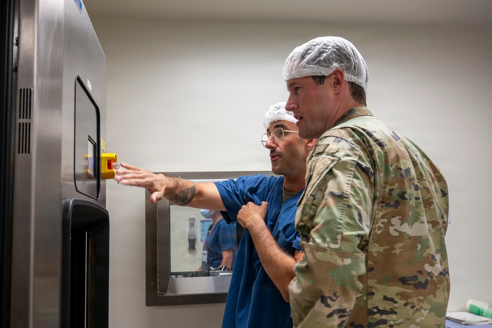Two individuals wearing hairnets are examining a large stainless steel medical or industrial equipment unit with a control panel and viewing window. One person is dressed in blue scrubs and glasses, pointing at the control panel, while the other wears military camouflage (OCP/MultiCam pattern) uniform. The setting appears to be a sterile or controlled environment such as a medical facility or laboratory.