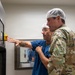 Two individuals wearing hairnets are examining a large stainless steel medical or industrial equipment unit with a control panel and viewing window. One person is dressed in blue scrubs and glasses, pointing at the control panel, while the other wears military camouflage (OCP/MultiCam pattern) uniform. The setting appears to be a sterile or controlled environment such as a medical facility or laboratory.