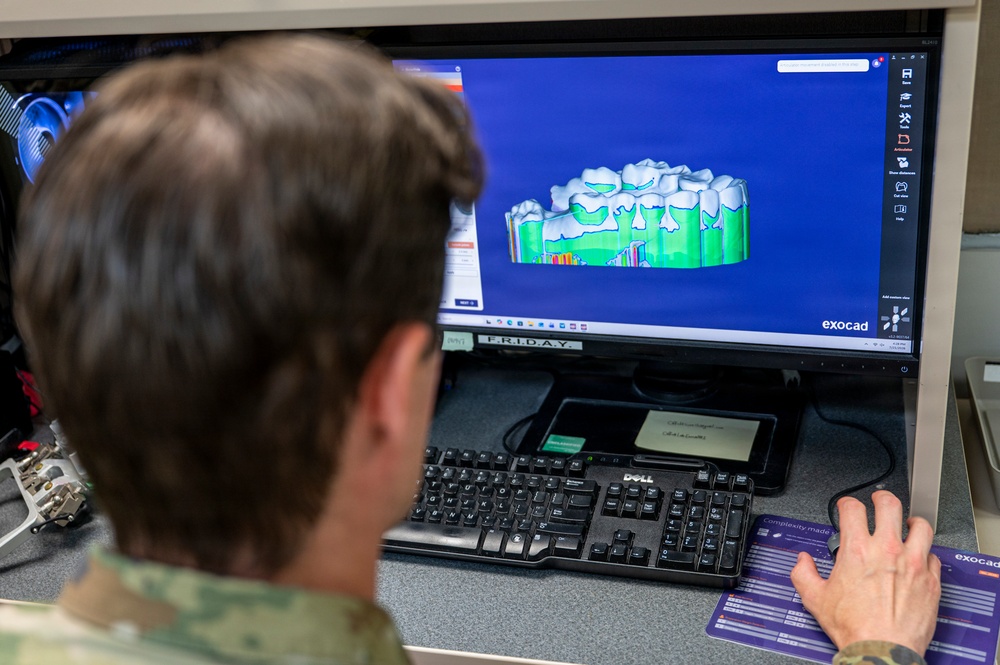 A person wearing military camouflage uniform is working at a desktop computer workstation running Exocad dental CAD software. The monitor displays a 3D model of dental crowns or a dental bridge rendered in green and white on a blue background. The setting appears to be a military dental laboratory environment.