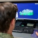 A person wearing military camouflage uniform is working at a desktop computer workstation running Exocad dental CAD software. The monitor displays a 3D model of dental crowns or a dental bridge rendered in green and white on a blue background. The setting appears to be a military dental laboratory environment.