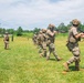 A group of military personnel in full combat gear are conducting a field training exercise on a grassy range. Soldiers are moving in a line formation while carrying rifles in various ready positions, suggesting a tactical movement or shooting drill. The setting appears to be a military training facility with a cleared grass field flanked by pine trees.