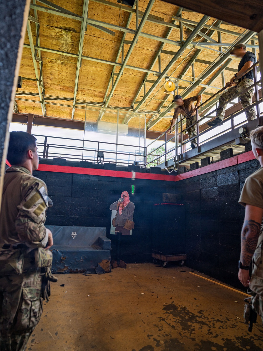 Military or law enforcement personnel are conducting a close-quarters battle (CQB) training exercise inside what appears to be a purpose-built shoot house or tactical training facility. Personnel in camouflage uniforms are positioned on both the ground floor and an elevated second-floor catwalk/mezzanine level. A cardboard silhouette target depicting a masked figure is visible on the ground floor as a training prop.