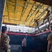 Military or law enforcement personnel are conducting a close-quarters battle (CQB) training exercise inside what appears to be a purpose-built shoot house or tactical training facility. Personnel in camouflage uniforms are positioned on both the ground floor and an elevated second-floor catwalk/mezzanine level. A cardboard silhouette target depicting a masked figure is visible on the ground floor as a training prop.