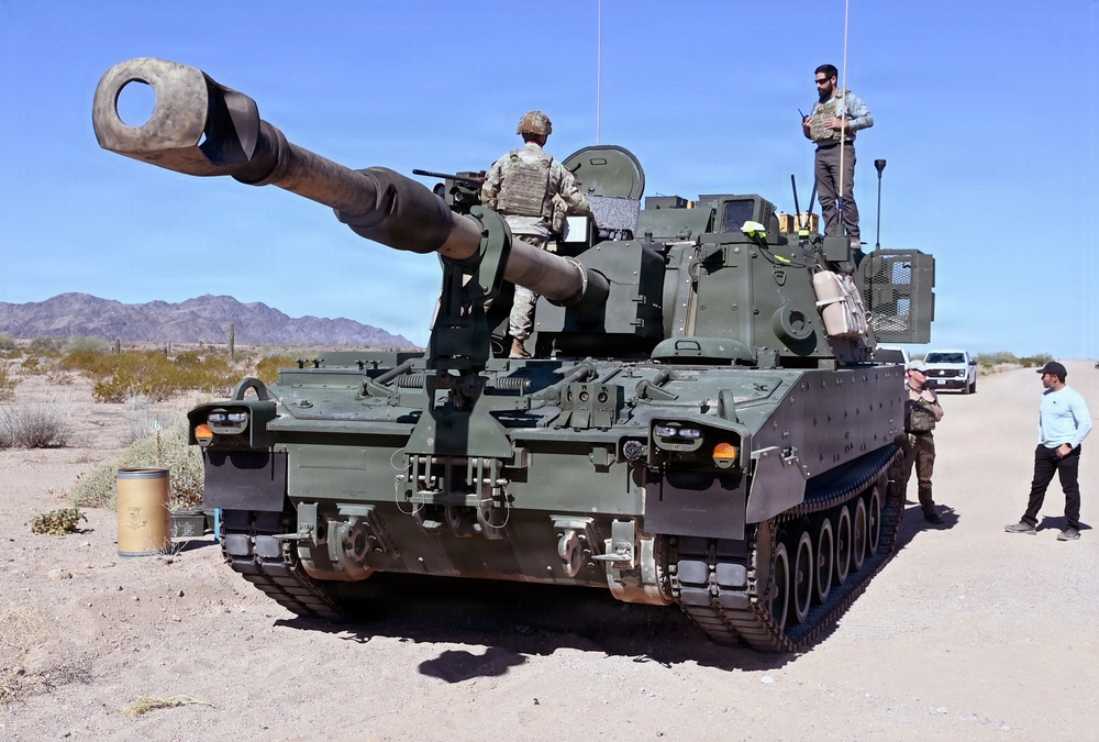 A self-propelled artillery vehicle, identified as an M109 Paladin howitzer, is positioned in an arid desert environment with mountains visible in the background. Several personnel, including uniformed soldiers wearing body armor and civilians, are gathered around and on top of the vehicle. The setting appears to be a training exercise or demonstration at a desert testing or training range.