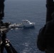 The image is taken from inside a military helicopter or aircraft, showing an aerial view of a white patrol or coast guard vessel on open ocean water. Personnel in high-visibility vests are visible on the deck of the vessel, and a smaller rigid inflatable boat (RIB) appears to be alongside it. In the foreground, a military weapon (likely a door-mounted machine gun) and a silhouetted crew member are visible, indicating this is a military aerial observation or overwatch operation.