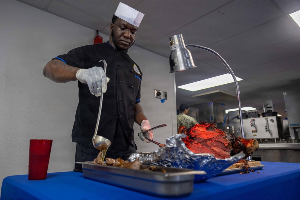 A chef wearing a black uniform and white paper toque is carving or serving a large roasted meat (likely a whole roasted pig or large roast) on a foil-lined tray under a heat lamp. The setting appears to be a military or institutional kitchen/dining facility, with another person visible in the background. The chef is using a ladle and tongs/fork while wearing white latex gloves.