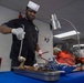 A chef wearing a black uniform and white paper toque is carving or serving a large roasted meat (likely a whole roasted pig or large roast) on a foil-lined tray under a heat lamp. The setting appears to be a military or institutional kitchen/dining facility, with another person visible in the background. The chef is using a ladle and tongs/fork while wearing white latex gloves.