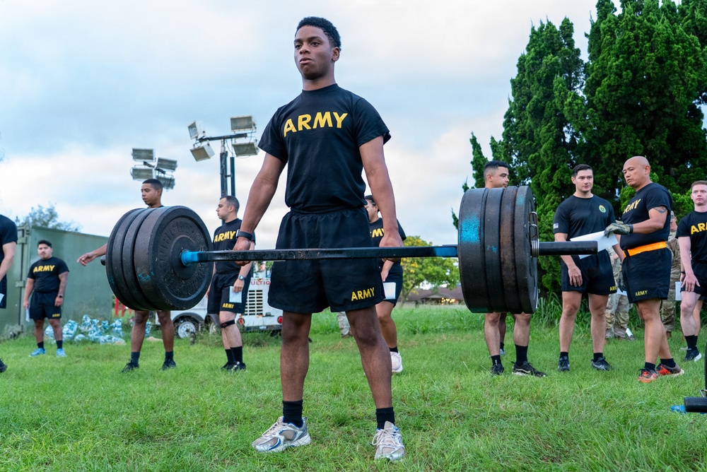A U.S. Army soldier performs a deadlift with a heavily loaded barbell on a grass field during what appears to be a physical fitness assessment or competition. Multiple other soldiers in matching black Army PT (Physical Training) uniforms observe in the background. The setting appears to be an outdoor military installation during overcast daylight conditions.