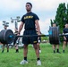 A U.S. Army soldier performs a deadlift with a heavily loaded barbell on a grass field during what appears to be a physical fitness assessment or competition. Multiple other soldiers in matching black Army PT (Physical Training) uniforms observe in the background. The setting appears to be an outdoor military installation during overcast daylight conditions.