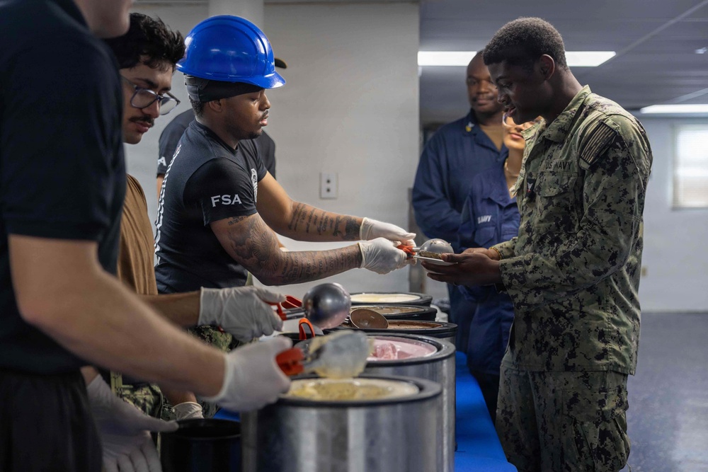 A food service activity is taking place in what appears to be a military facility, where individuals wearing 'FSA' (Food Service Attendant) shirts and gloves are serving food to uniformed U.S. Navy personnel. The server in the foreground is wearing a blue hard hat and black FSA shirt while scooping food from large metal containers. At least one person in U.S. Navy digital camouflage (NWU) uniform is receiving food, with others in blue Navy working uniforms visible in the background.