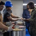 A food service activity is taking place in what appears to be a military facility, where individuals wearing 'FSA' (Food Service Attendant) shirts and gloves are serving food to uniformed U.S. Navy personnel. The server in the foreground is wearing a blue hard hat and black FSA shirt while scooping food from large metal containers. At least one person in U.S. Navy digital camouflage (NWU) uniform is receiving food, with others in blue Navy working uniforms visible in the background.