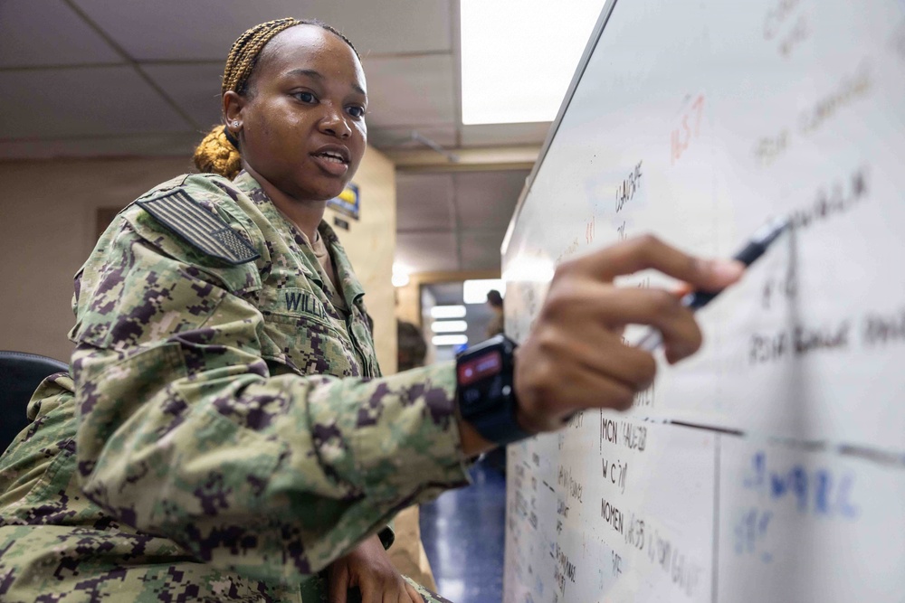 A female U.S. Navy service member wearing NWU Type III (woodland digital camouflage) uniform is writing or pointing on a whiteboard. Her name tape reads 'WILLIAMS' and she is wearing a smartwatch on her left wrist. The setting appears to be a military office or operations room with text and data visible on the whiteboard.