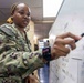 A female U.S. Navy service member wearing NWU Type III (woodland digital camouflage) uniform is writing or pointing on a whiteboard. Her name tape reads 'WILLIAMS' and she is wearing a smartwatch on her left wrist. The setting appears to be a military office or operations room with text and data visible on the whiteboard.