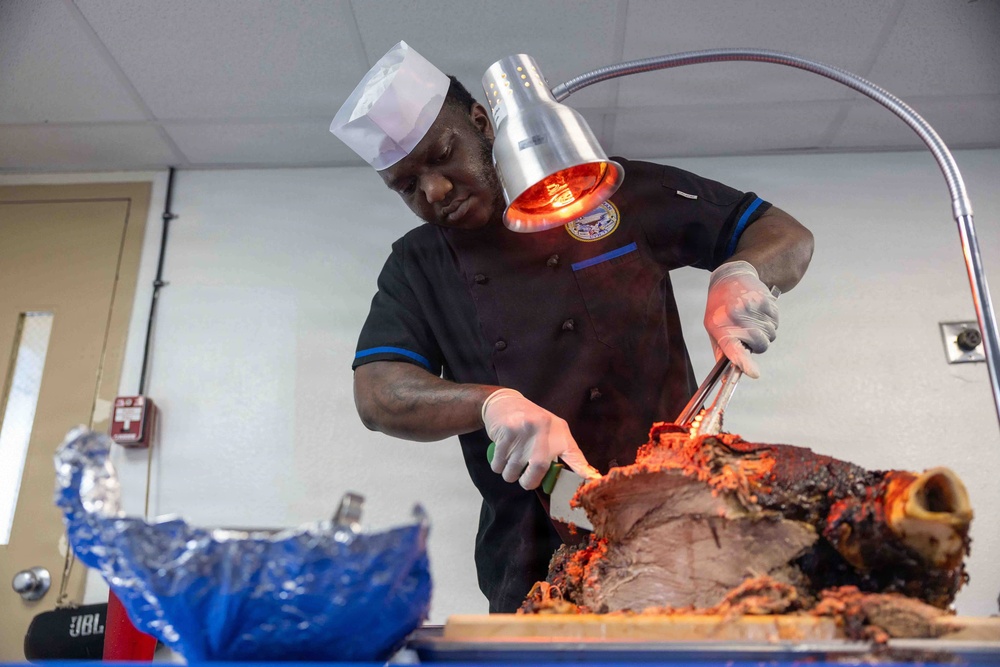 A chef in a black uniform and white toque is carving a large roasted meat joint, likely a leg of lamb or pork, using a knife and tongs. A heat lamp positioned overhead keeps the meat warm during service. The setting appears to be a food service or catering environment.