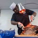 A chef in a black uniform and white toque is carving a large roasted meat joint, likely a leg of lamb or pork, using a knife and tongs. A heat lamp positioned overhead keeps the meat warm during service. The setting appears to be a food service or catering environment.