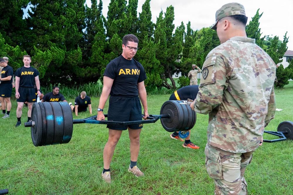 U.S. Army soldiers are conducting an outdoor physical fitness training session on a grass field. A soldier in PT (physical training) uniform is performing a hex bar/trap bar deadlift while a uniformed soldier in OCP (Operational Camouflage Pattern) observes and evaluates. Additional soldiers in black Army PT uniforms are visible in the background performing various exercises.
