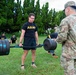 U.S. Army soldiers are conducting an outdoor physical fitness training session on a grass field. A soldier in PT (physical training) uniform is performing a hex bar/trap bar deadlift while a uniformed soldier in OCP (Operational Camouflage Pattern) observes and evaluates. Additional soldiers in black Army PT uniforms are visible in the background performing various exercises.