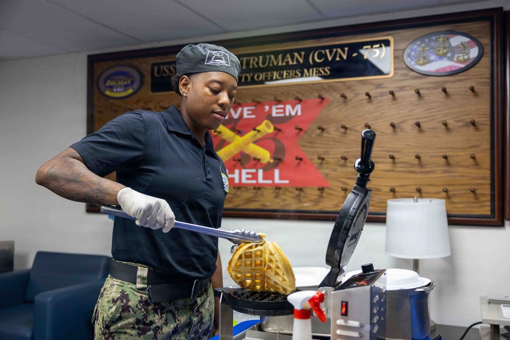 A U.S. Navy service member in uniform is preparing waffles using a commercial waffle iron in what appears to be the Chief Petty Officers Mess aboard the USS Harry S. Truman (CVN-75). The sailor is wearing Navy Working Uniform (NWU) camouflage pants, a dark polo shirt, a gray cap, and white food-service gloves. The setting is a naval vessel's dining facility, identifiable by the prominent ship placard on the wall.