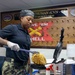 A U.S. Navy service member in uniform is preparing waffles using a commercial waffle iron in what appears to be the Chief Petty Officers Mess aboard the USS Harry S. Truman (CVN-75). The sailor is wearing Navy Working Uniform (NWU) camouflage pants, a dark polo shirt, a gray cap, and white food-service gloves. The setting is a naval vessel's dining facility, identifiable by the prominent ship placard on the wall.