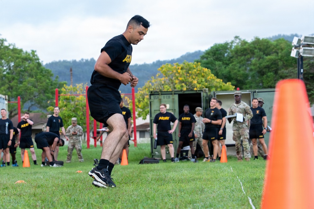 A U.S. Army soldier in black PT (Physical Training) uniform is running on a grass field during what appears to be a physical fitness test or assessment. Multiple other soldiers and personnel in both black Army PT uniforms and camouflage ACU uniforms are visible in the background observing or waiting their turn. Orange traffic cones mark the course boundaries, and the setting appears to be a military installation with trees and hills visible in the background.