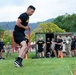 A U.S. Army soldier in black PT (Physical Training) uniform is running on a grass field during what appears to be a physical fitness test or assessment. Multiple other soldiers and personnel in both black Army PT uniforms and camouflage ACU uniforms are visible in the background observing or waiting their turn. Orange traffic cones mark the course boundaries, and the setting appears to be a military installation with trees and hills visible in the background.