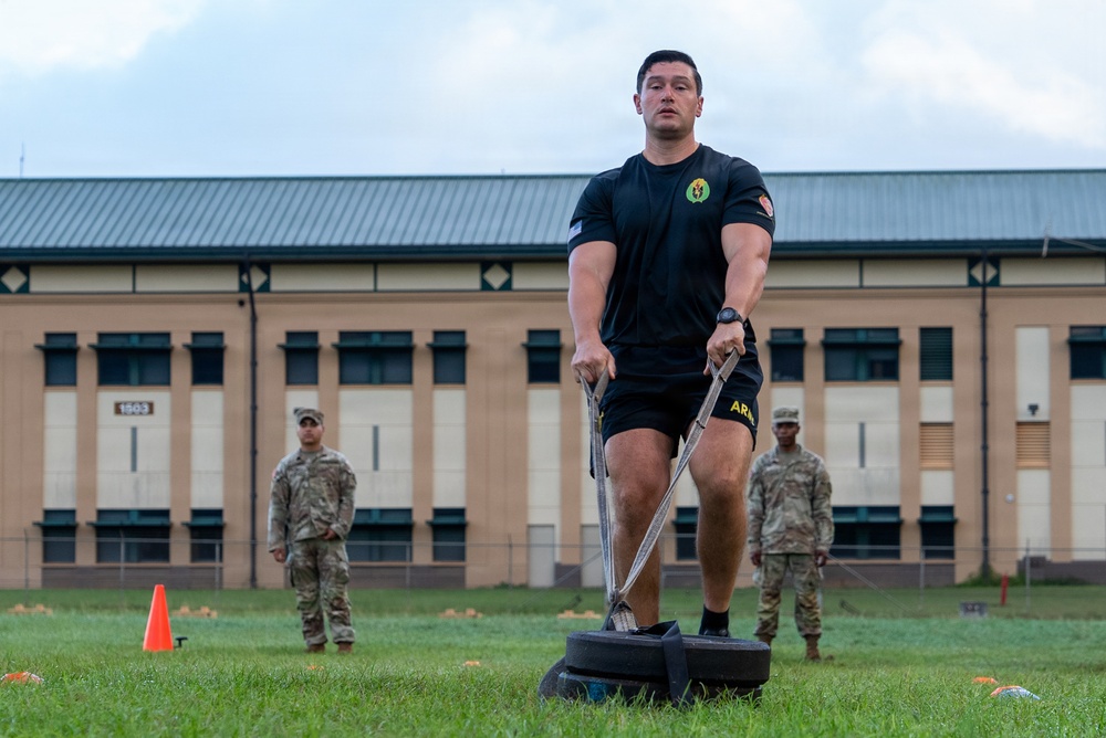 A muscular soldier in black Army PT (Physical Training) uniform is performing a sled drag exercise on a grass field at what appears to be a military installation. He is pulling a weighted sled using straps while two other soldiers in camouflage uniforms observe in the background. The setting appears to be an Army Combat Fitness Test (ACFT) or physical training event.
