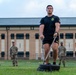 A muscular soldier in black Army PT (Physical Training) uniform is performing a sled drag exercise on a grass field at what appears to be a military installation. He is pulling a weighted sled using straps while two other soldiers in camouflage uniforms observe in the background. The setting appears to be an Army Combat Fitness Test (ACFT) or physical training event.