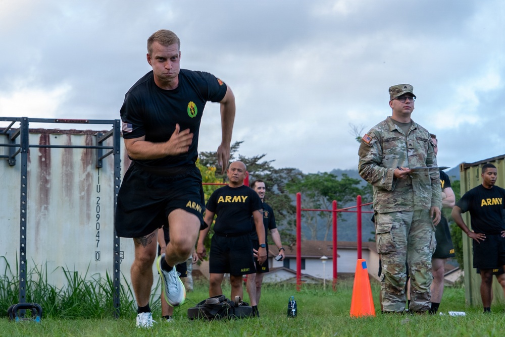 Soldados del ejército estadounidense están realizando una prueba de aptitud física o un ejercicio de entrenamiento en un campo de césped. Un soldado con uniforme negro de PT (Entrenamiento físico) del ejército corre pasando un cono naranja mientras un soldado uniformado con OCP (Patrón de camuflaje operativo) sostiene un portapapeles, probablemente anotando o cronometrando el evento. Varios otros soldados con equipo PT negro del ejército observan al fondo.