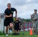 U.S. Army soldiers are conducting a physical fitness test or training exercise on a grass field. A soldier in black Army PT (Physical Training) uniform is sprinting past an orange cone while a uniformed soldier in OCP (Operational Camouflage Pattern) holds a clipboard, likely scoring or timing the event. Several other soldiers in black Army PT gear observe in the background.