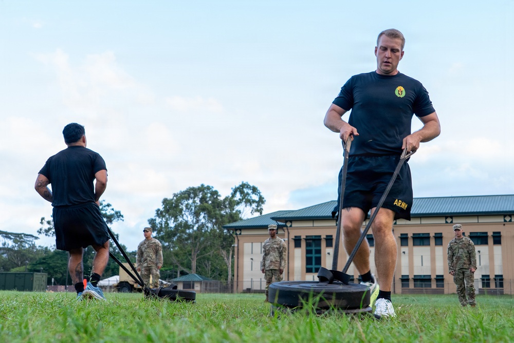U.S. Army soldiers are conducting physical training on a grass field at what appears to be a military installation. The foreground soldier is performing a sled drag exercise, pulling a weighted sled using straps, while another soldier performs a similar exercise in the background. Several uniformed soldiers in camouflage ACUs observe or supervise the training session.