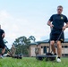 U.S. Army soldiers are conducting physical training on a grass field at what appears to be a military installation. The foreground soldier is performing a sled drag exercise, pulling a weighted sled using straps, while another soldier performs a similar exercise in the background. Several uniformed soldiers in camouflage ACUs observe or supervise the training session.